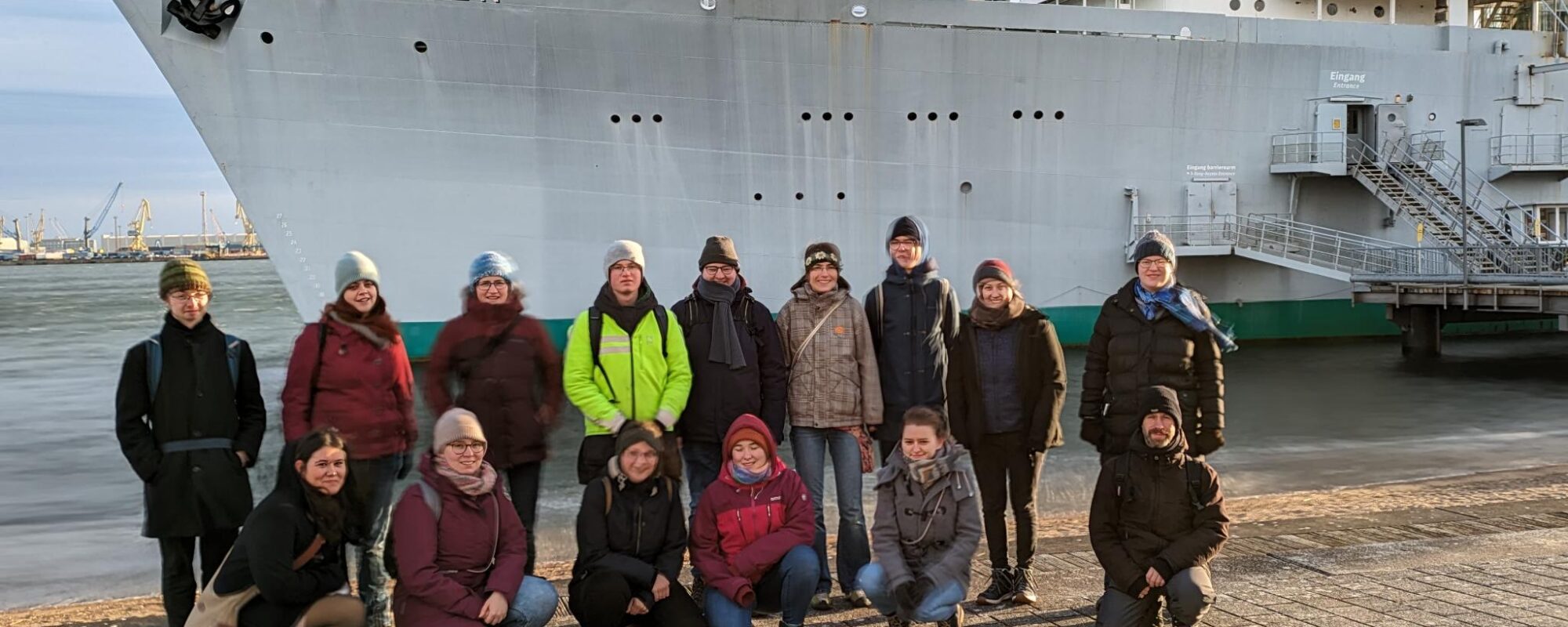 Die Gruppe Studierender steht vor einem Schiff an der Ostsee.